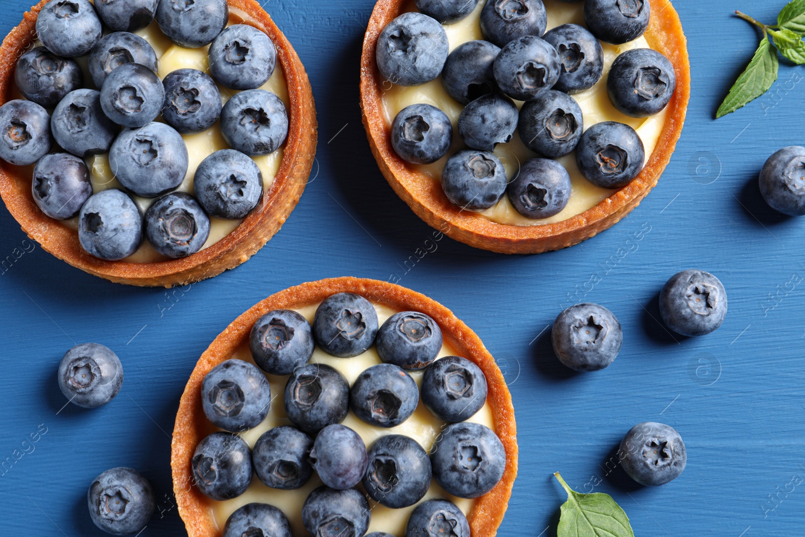 Photo of Tartlets with fresh blueberries on blue wooden table, flat lay. Delicious dessert