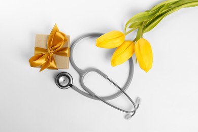 Photo of Stethoscope, gift box and yellow tulips on white background, flat lay. Happy Doctor's Day