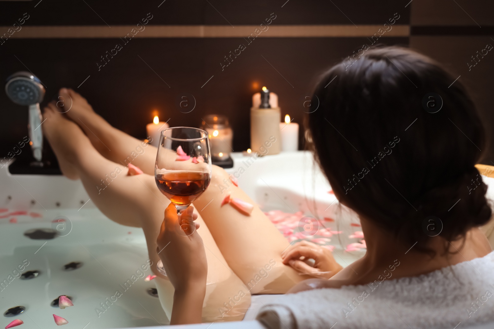 Photo of Woman holding glass of wine while taking bath with rose petals, back view. Romantic atmosphere