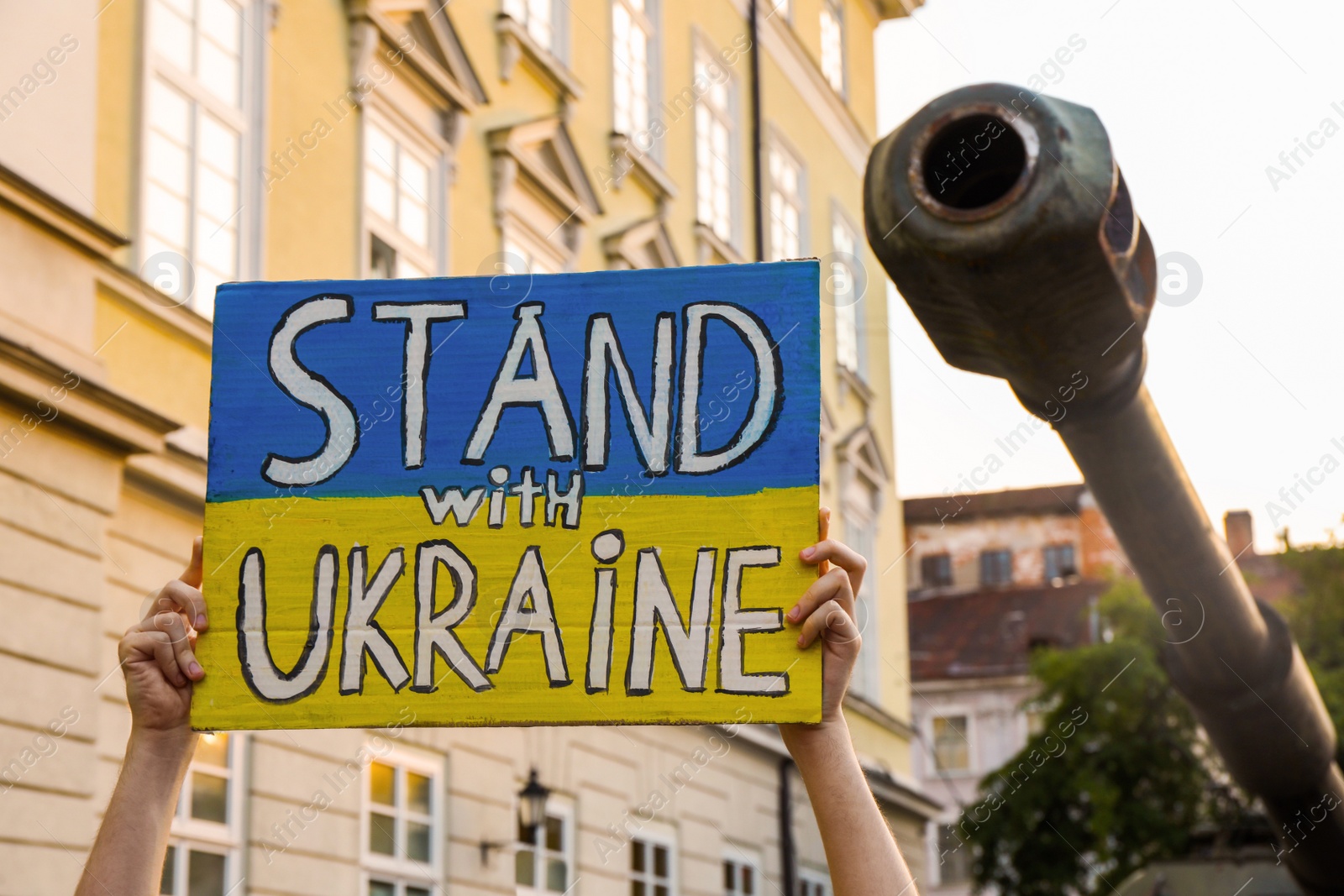 Photo of Woman holding poster in colors of national flag and words Stand With Ukraine near broken military tank outdoors, closeup