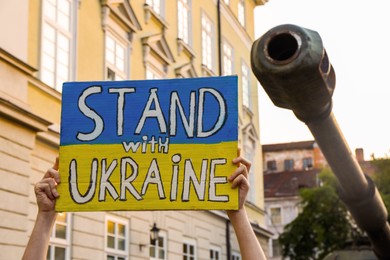Photo of Woman holding poster in colors of national flag and words Stand With Ukraine near broken military tank outdoors, closeup