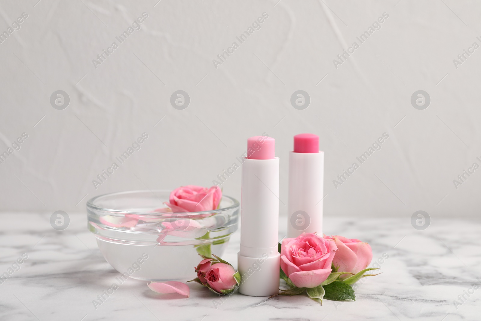 Photo of Lip balms, bowl with water and roses on white marble table, space for text