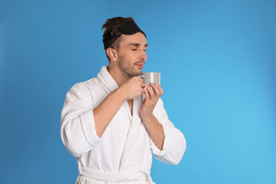 Young man in bathrobe with cup of coffee on light blue background