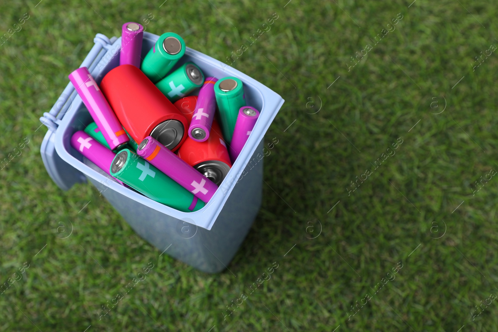 Photo of Many used batteries in recycling bin on green grass, above view. Space for text