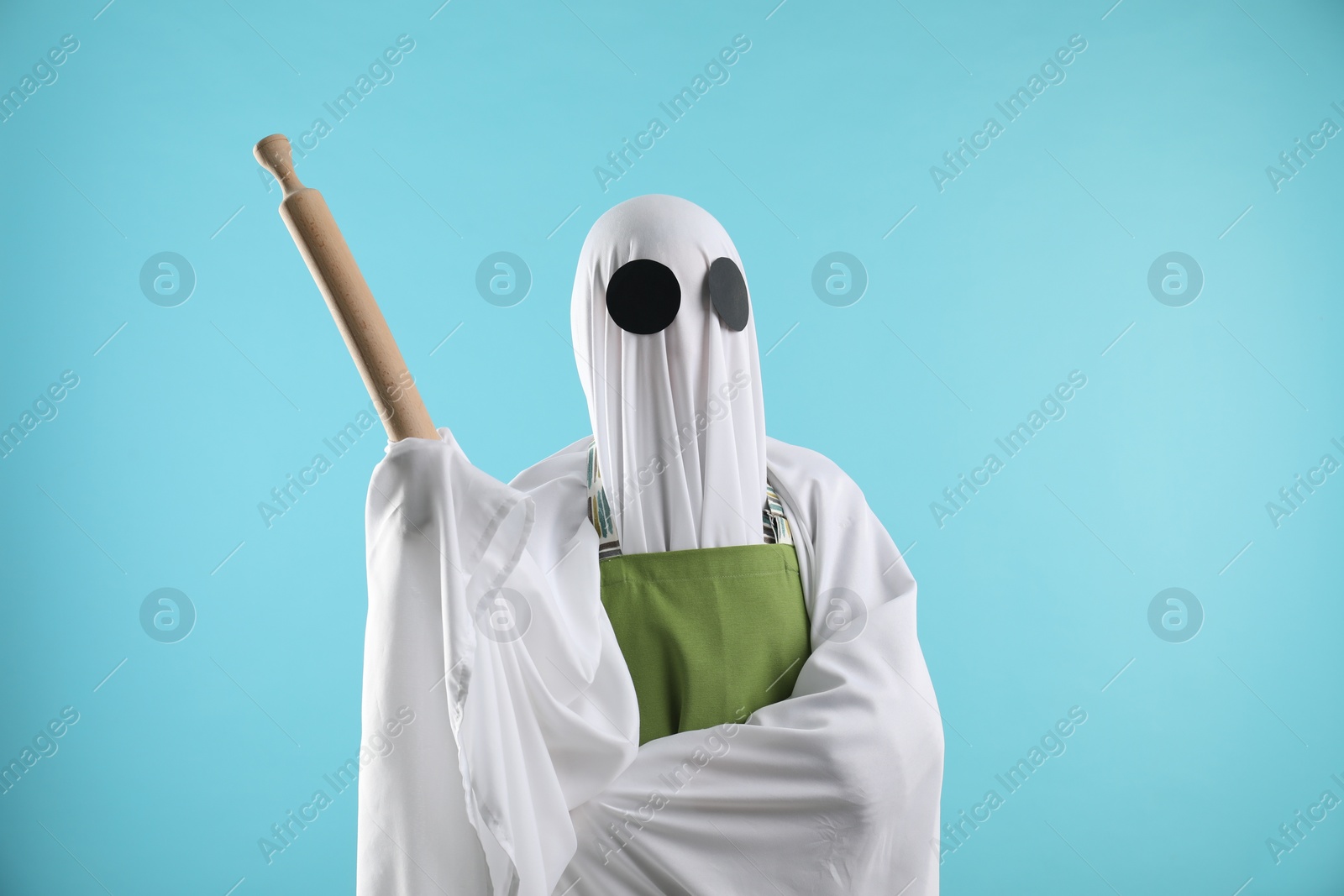 Photo of Woman in ghost costume and apron with rolling pin on light blue background