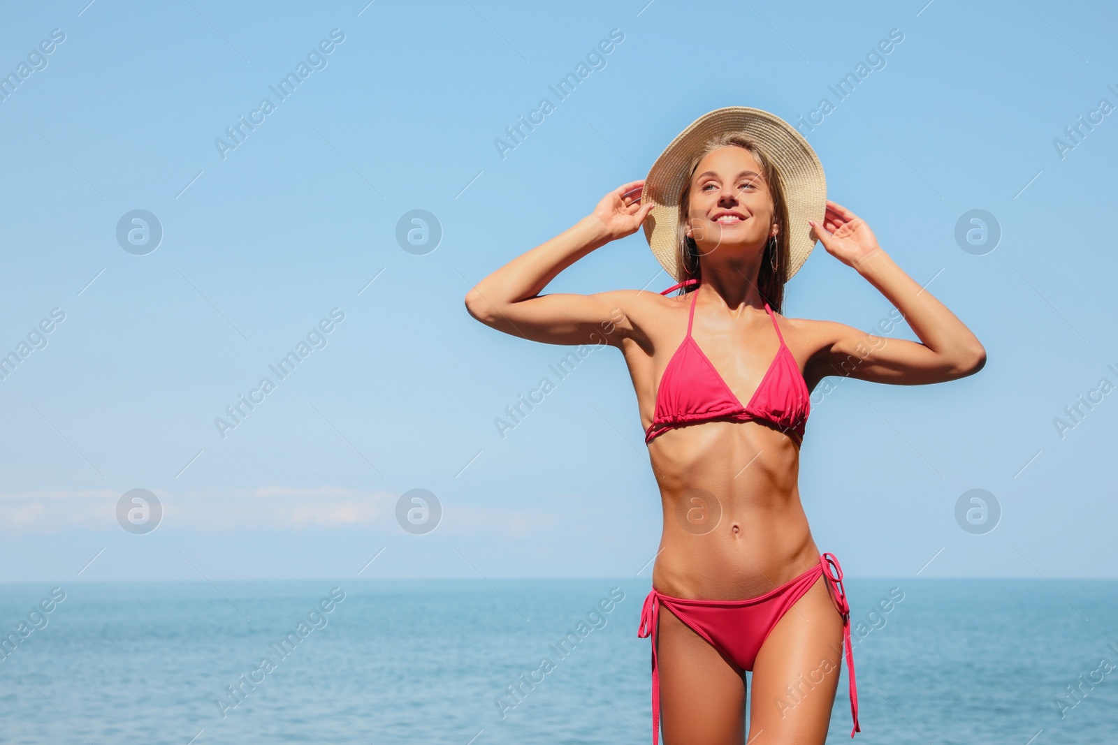 Photo of Beautiful young woman in stylish bikini and straw hat on seashore, space for text