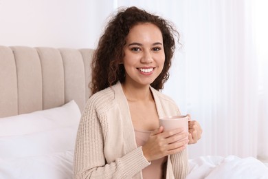 Happy African American woman with cup of drink in bed at home