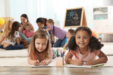 Photo of Cute girls drawing on floor while kindergarten teacher reading book to other children indoors