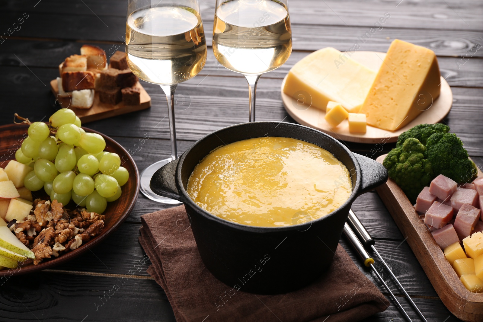 Photo of Fondue pot with melted cheese, glasses of wine and different products on black wooden table