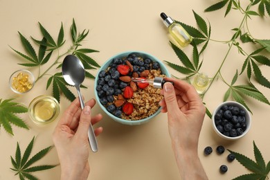 Photo of Top view of woman dripping THC tincture or CBD oil into oatmeal bowl on beige background, closeup