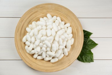 Silk cocoons with plate and mulberry leaves on white wooden table, flat lay