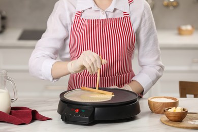 Photo of Woman cooking delicious crepe on electric pancake maker at white marble table in kitchen, closeup