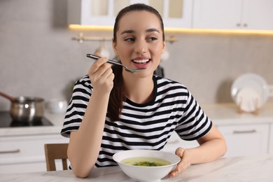Beautiful woman eating tasty soup at white table in kitchen