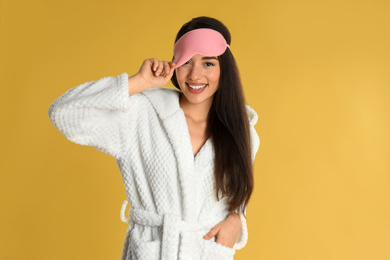 Beautiful young woman in bathrobe and eye sleeping mask on yellow background