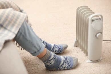 Photo of Young woman warming feet near electric heater at home, closeup
