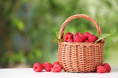 Photo of Wicker basket with tasty ripe raspberries and leaves on white table against blurred green background, space for text
