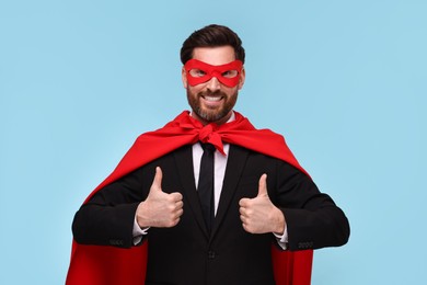 Businessman in red superhero cape and mask showing thumbs up on light blue background