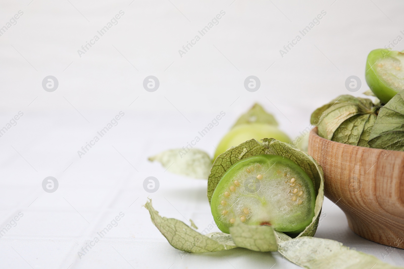 Photo of Fresh green tomatillos with husk in bowl on white tiled table, closeup. Space for text