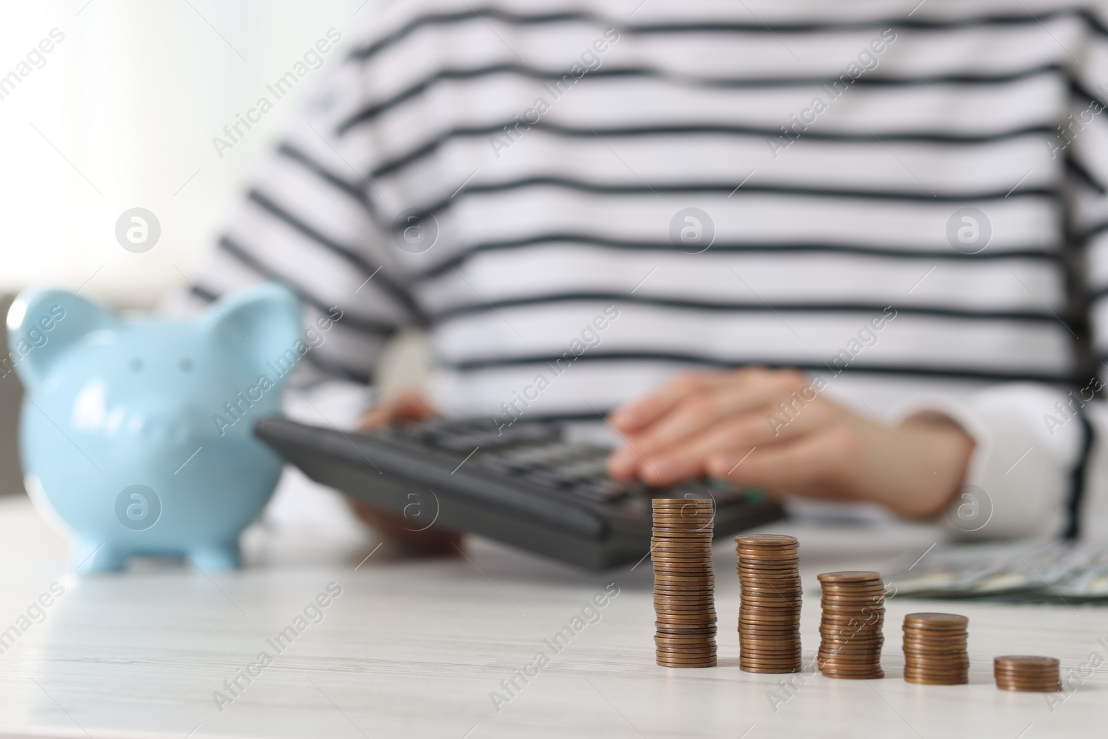 Photo of Financial savings. Woman using calculator at white wooden table, focus stacked coins