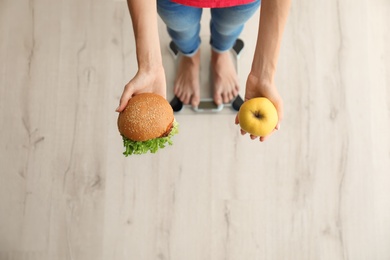 Photo of Woman holding tasty sandwich and fresh apple while measuring her weight on floor scales, top view. Choice between diet and unhealthy food