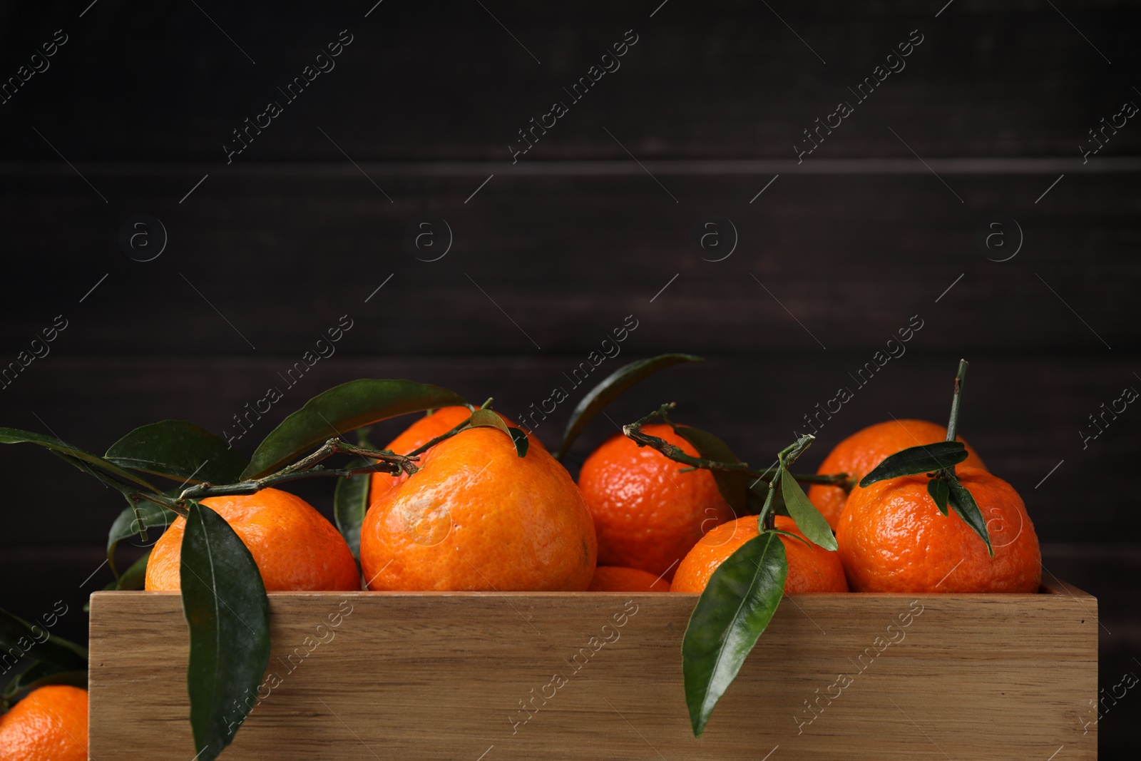 Photo of Fresh ripe tangerines with green leaves in wooden crate on dark background, closeup. Space for text