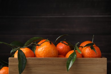 Photo of Fresh ripe tangerines with green leaves in wooden crate on dark background, closeup. Space for text