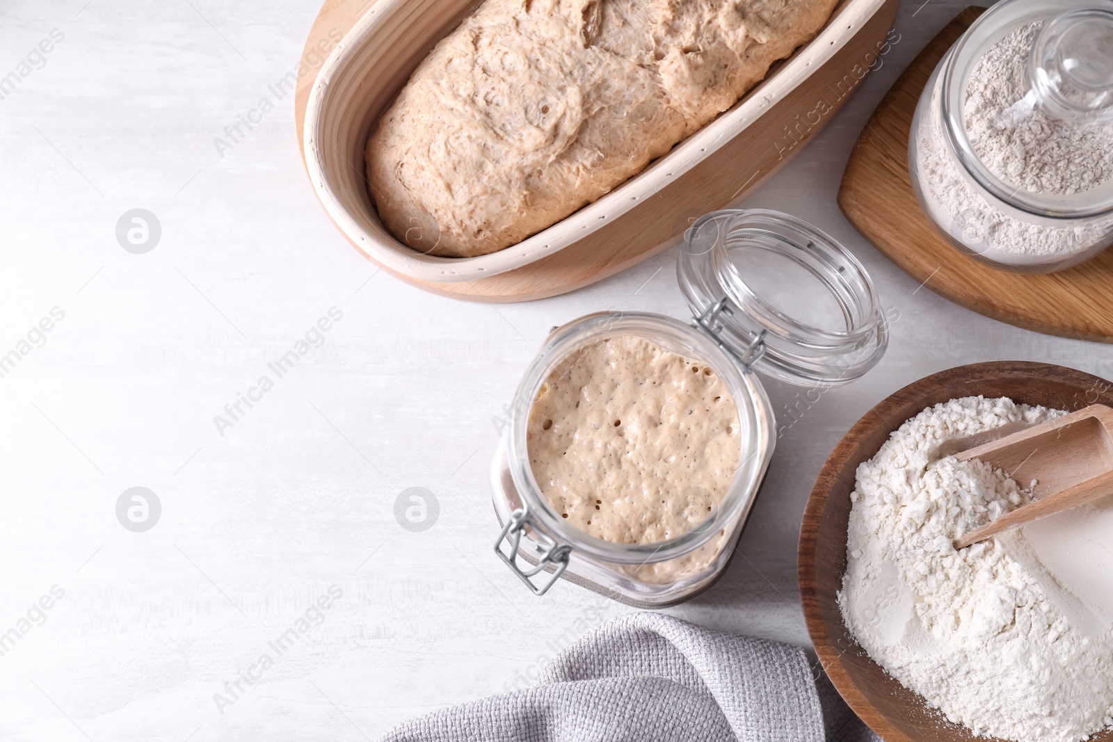 Photo of Sourdough starter in glass jar, flour and dough on light table, flat lay. Space for text