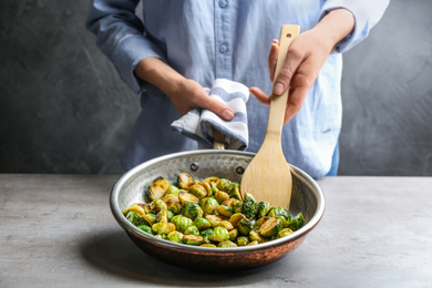 Photo of Woman with frying pan of roasted Brussels sprouts at grey table, closeup