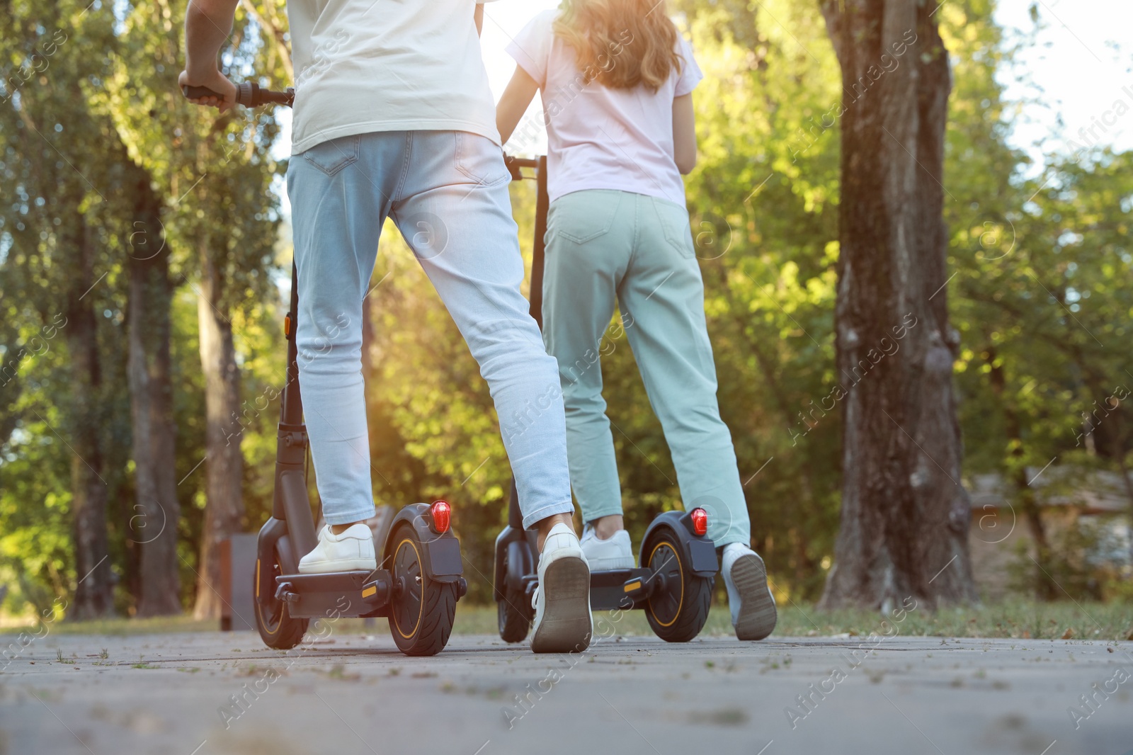 Photo of Couple riding modern electric kick scooters in park, back view