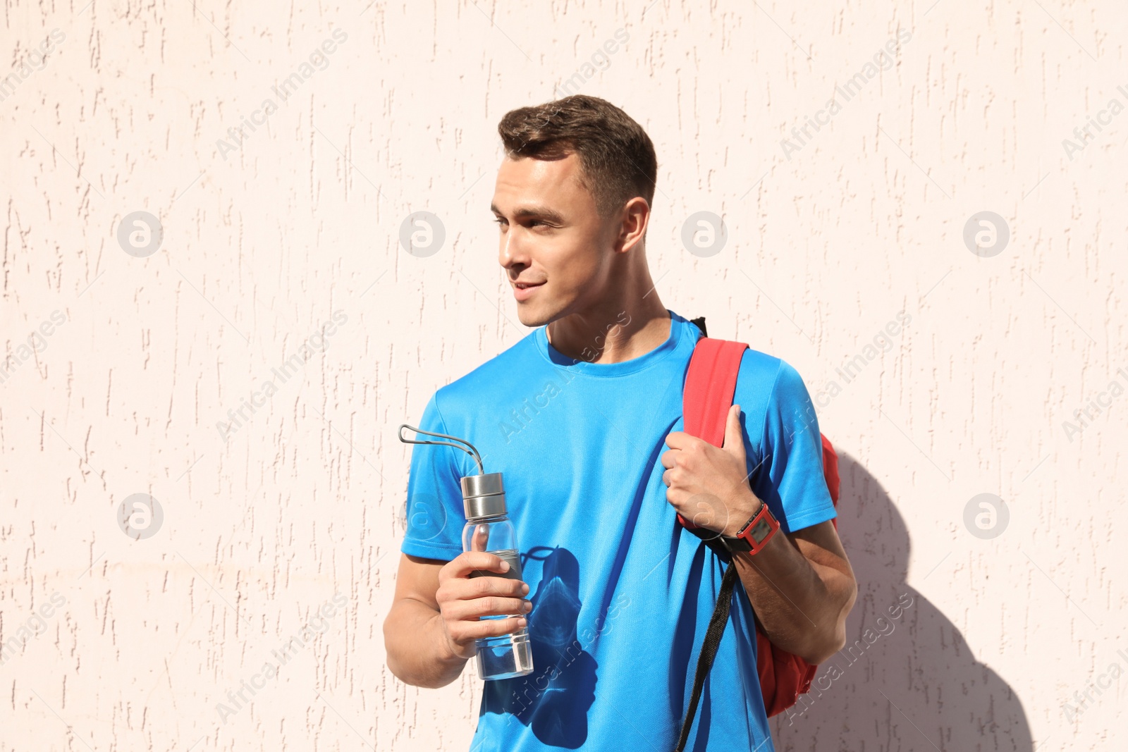 Photo of Young sporty man with backpack holding bottle of water near wall outdoors on sunny day