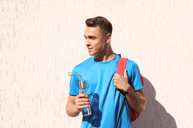 Photo of Young sporty man with backpack holding bottle of water near wall outdoors on sunny day