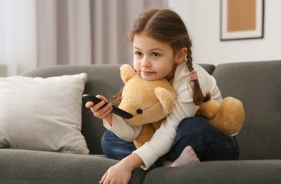 Photo of Little girl changing TV channels with remote control on sofa at home