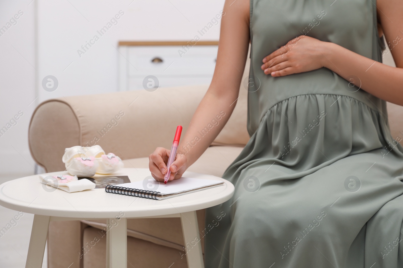 Photo of Pregnant woman with baby's accessories writing names list at small white table, closeup