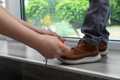 Mother helping son to tie shoe laces near window at home, closeup