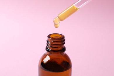 Photo of Dripping cosmetic oil from pipette into bottle on pink background, closeup