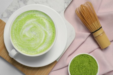 Cup of fresh matcha latte, powder and bamboo whisk on white table, flat lay