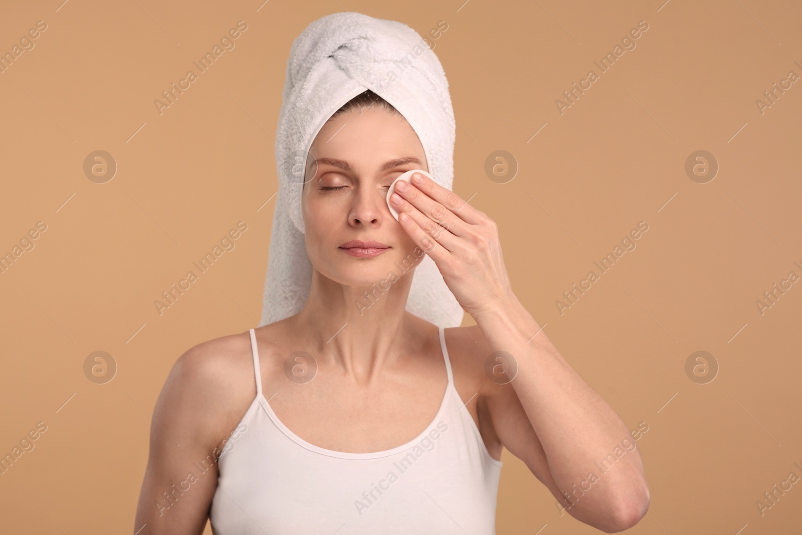 Photo of Beautiful woman in terry towel removing makeup with cotton pad on beige background