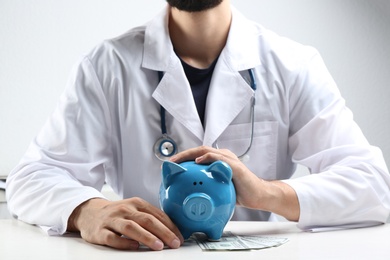 Photo of Doctor with piggybank and money at white table indoors, closeup. Medical insurance concept