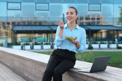 Photo of Smiling businesswoman eating from lunch box near laptop outdoors. Space for text