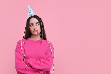 Sad woman in party hat with blower and streamers on pink background, space for text