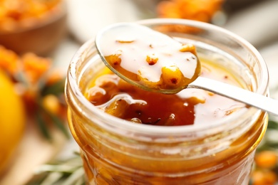 Photo of Spoon with delicious sea buckthorn jam over jar, closeup