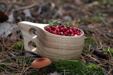 Photo of Many ripe lingonberries in wooden cup on ground near mushroom outdoors