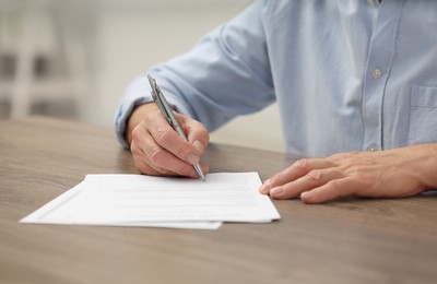 Photo of Senior man signing Last Will and Testament at wooden table, closeup