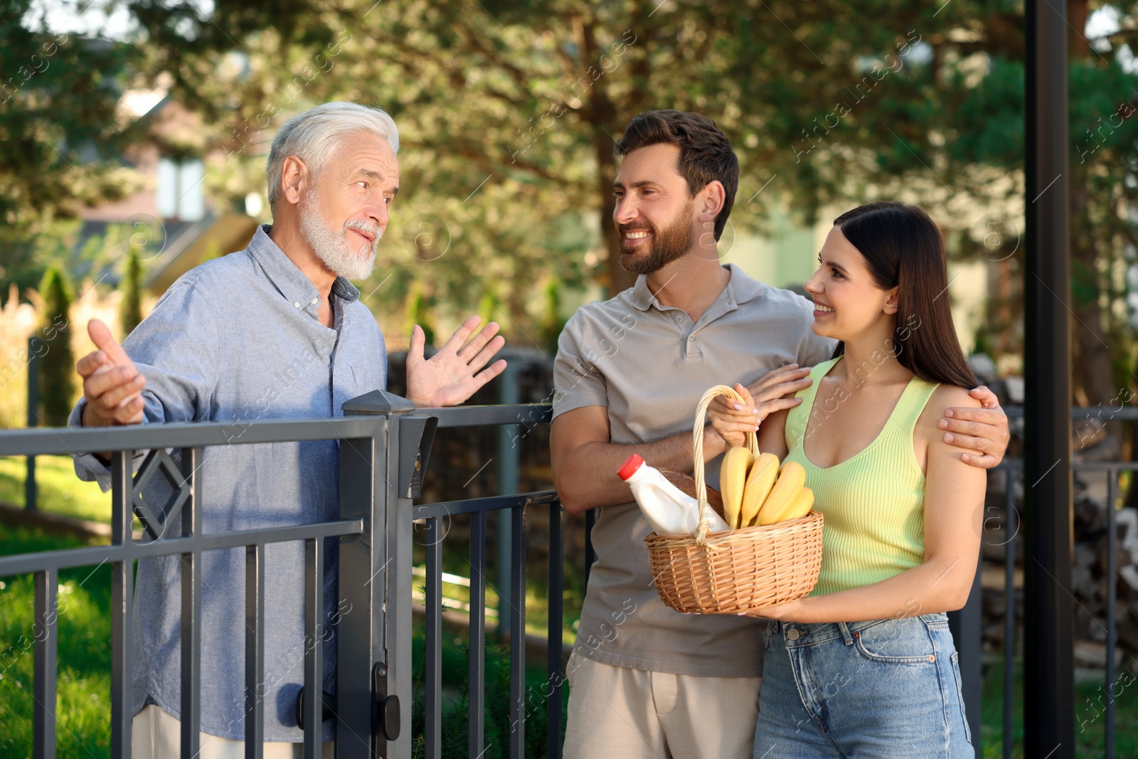 Photo of Friendly relationship with neighbours. Young couple with wicker basket of products treating senior man near fence outdoors