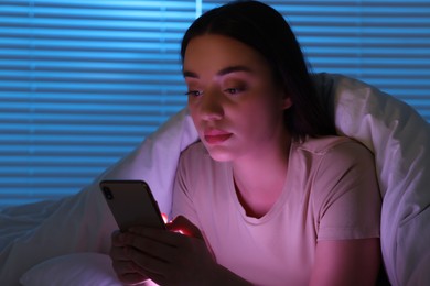 Photo of Young woman using modern smartphone in bed at night. Internet addiction