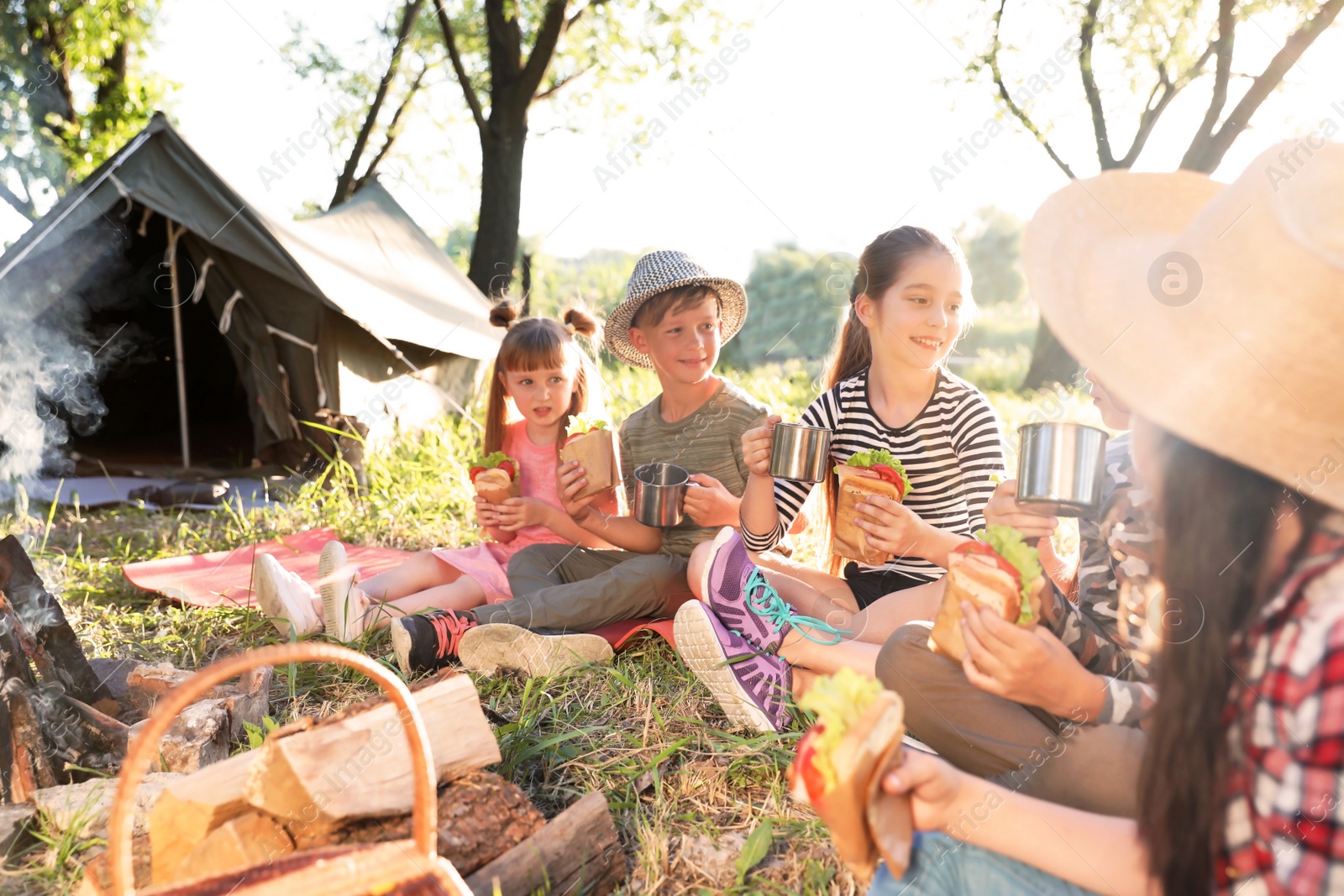 Photo of Little children eating sandwiches near bonfire and tent. Summer camp