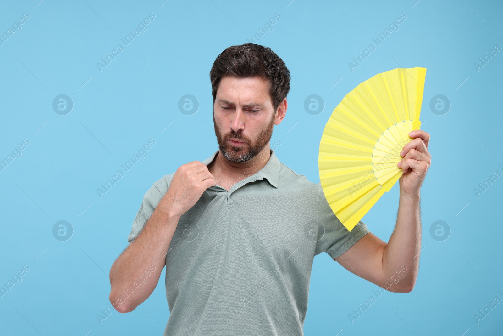 Photo of Unhappy man with hand fan suffering from heat on light blue background