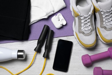 Photo of Smartphone, wireless earphones, clothes and sports equipment on white wooden table, flat lay. Personal training