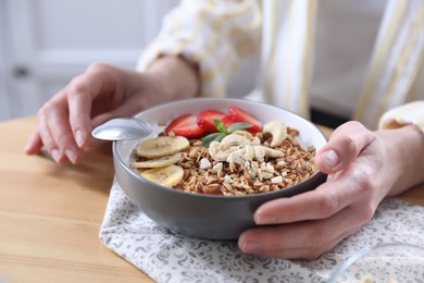 Photo of Woman eating tasty granola with banana, cashew and strawberries at wooden table indoors, closeup