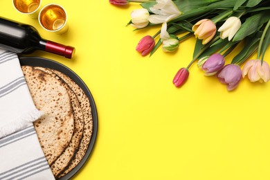 Photo of Tasty matzos, wine and fresh flowers on yellow background, space for text. Passover (Pesach) celebration